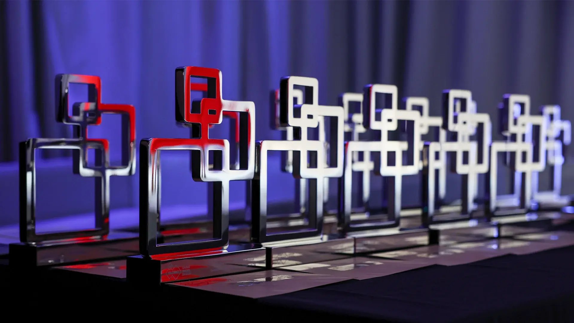 Row of modern geometric trophy awards displayed on a table under purple and blue stage lighting, ready for presentation at an event ceremony.