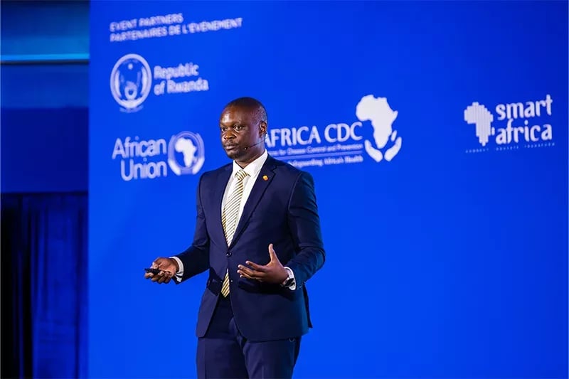A man in a dark suit with a yellow tie stands on stage, gesturing with his hands while speaking. The background displays the logos of the Republic of Rwanda, African Union, Africa CDC, and Smart Africa at an event in Kigali.