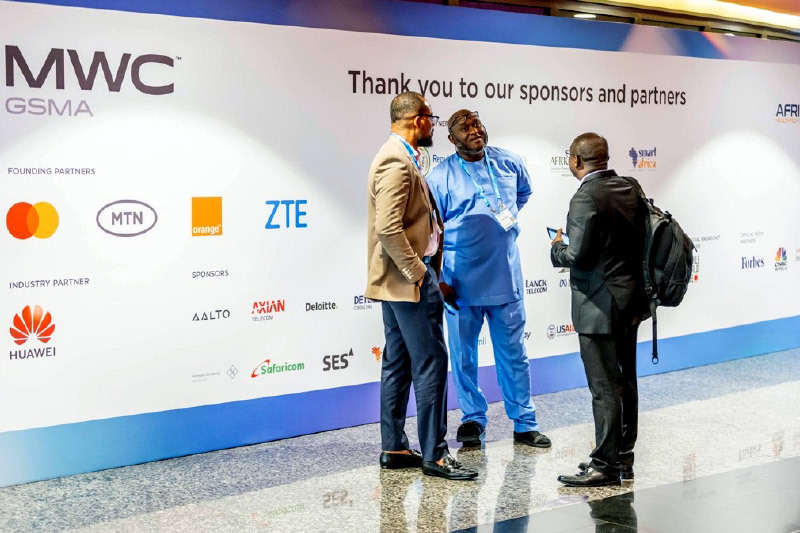 Three men converse in front of a large sponsor wall at MWC, displaying logos of partners such as Mastercard, MTN, Orange, ZTE, Huawei, and Deloitte.