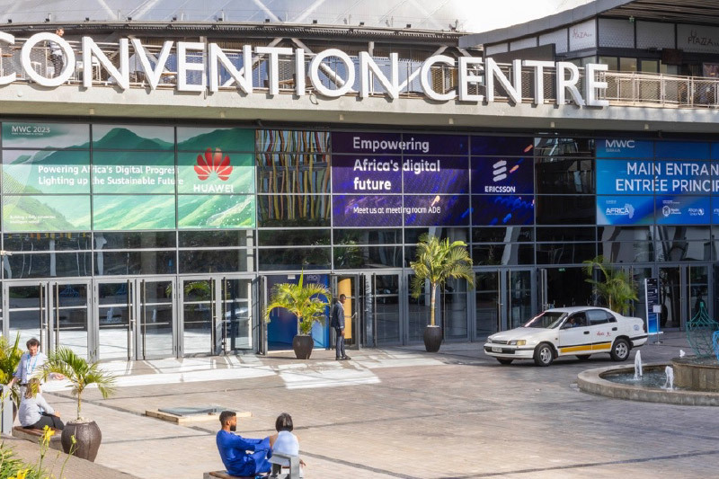 Exterior view of a convention centre with large banners for Huawei and Ericsson promoting digital progress in Africa, a white taxi parked in front, and people walking and sitting around the entrance area.
