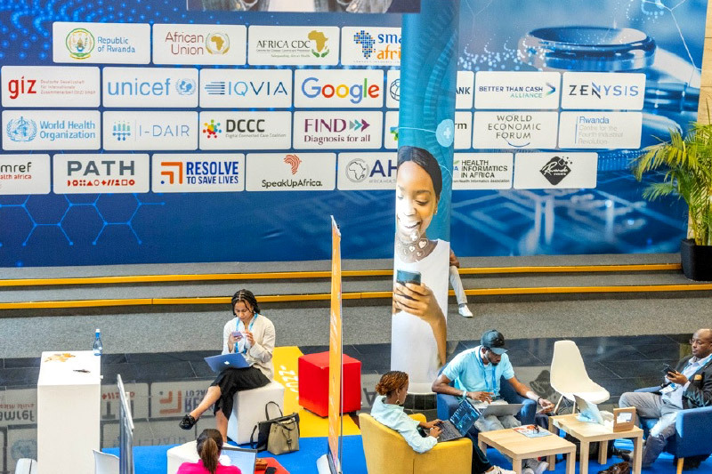 Conference attendees work and relax in a networking lounge area with sponsor logos displayed on a large backdrop, including organizations like Google, UNICEF, WHO, PATH, and the African Union, during a health and technology summit.